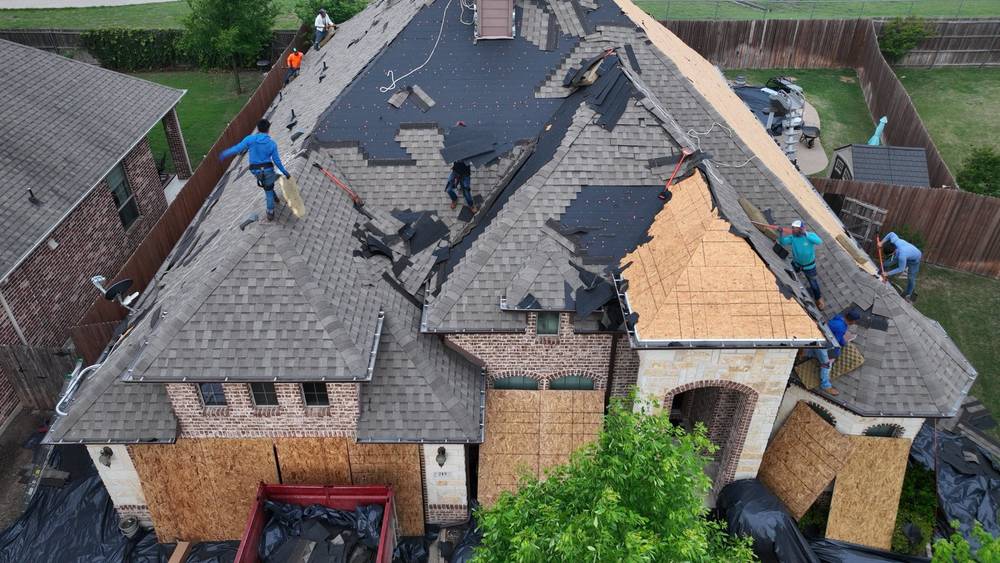 Roofing crew tearing storm-damaged shingles off a brick and stone home, dumpster staged below